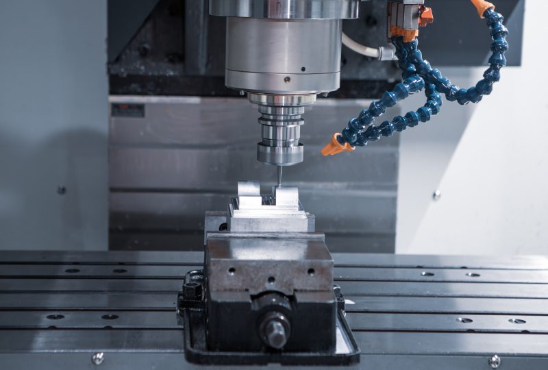 GettyImages-1866355504 A CNC machine operates on a metal piece secured in a vice, with coolant hoses directed at the machining area.